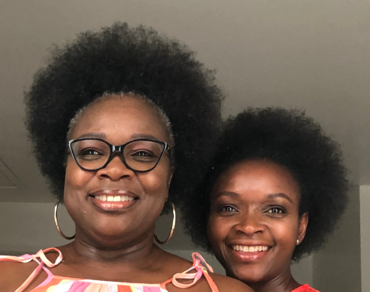Two smiling black women with large afro hair posing together. The woman on the left wears black cat-eye glasses, large gold hoop earrings, and a pink and white striped top. The women on the right is the Made With Nature Founder.