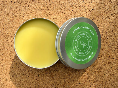 An open round metal tin of Made With Nature organic shea butter and sesame oil moisturiser, showing pale yellow balm inside, with the lid displaying a green label, set on a cork background.