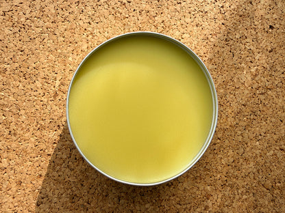 An open round metal tin of Made With Nature organic shea butter and sesame oil moisturiser, showing pale yellow balm inside, set on a cork background.