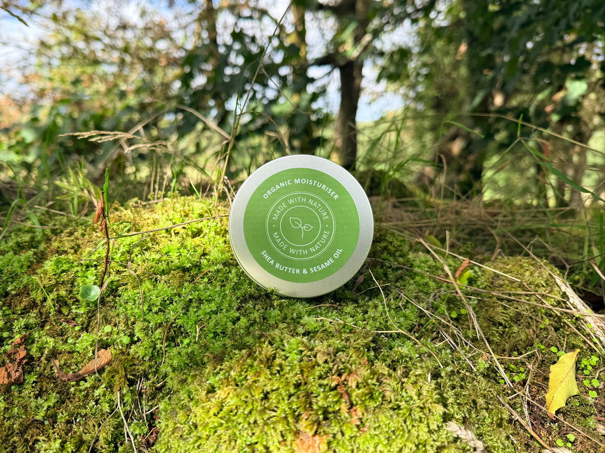 A round metal tin of Made With Nature organic shea butter and sesame oil moisturiser sits on a mossy forest ground with trees and blue sky in the background.